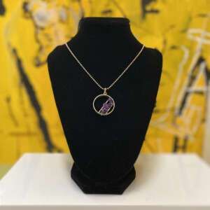 A silver necklace with a circular pendant featuring small purple stones is displayed on a black velvet stand. The background is a vibrant yellow with abstract patterns.