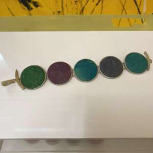 Five colorful round stones in a bracelet; green, purple, blue, dark purple, and turquoise. Set against a white surface with a yellow abstract background.