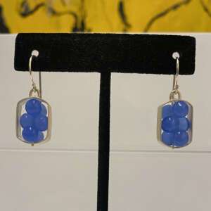 Silver earrings featuring vertically stacked blue beads, displayed on a black stand. Background includes a blurred yellow and black abstract.