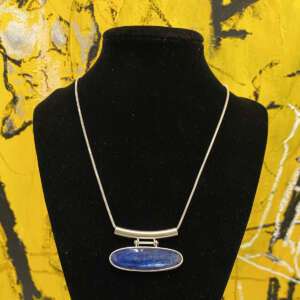 A silver necklace with a blue oval pendant displayed on a black stand. The background features an abstract yellow and black painted mural.