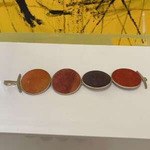 Close-up of a bracelet with four oval stones in earthy tones: orange, red, and purple. Set in silver, it rests on a white surface. Background is abstract yellow and black.