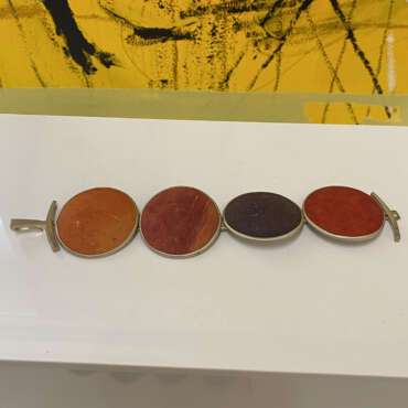 Close-up of a bracelet with four oval stones in earthy tones: orange, red, and purple. Set in silver, it rests on a white surface. Background is abstract yellow and black.